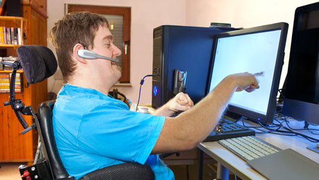 Mies istuu pyörätuolissa tietokoneella ja osoittaa näyttöä.