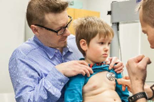 Lapsi istuu isän sylissä, lääkärin kuuntelee stetoskoopilla lapsen rintaa.