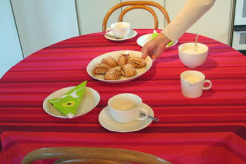 Käsi laittaa pöydälle pullavadin. Pöydällä on jo kahvikupit, sokeri ja maito.